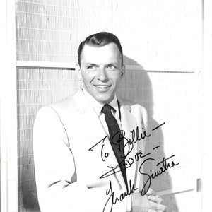 May include: Black and white photo of Frank Sinatra wearing a suit and tie. He is smiling and looking to the right. He has signed the photo "To Billie - Love - Frank Sinatra".