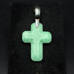 Könnte beinhalten: Ein hellgrüner Kreuzanhänger mit einer silberfarbenen Öse. Das Kreuz besteht aus einem glatten, polierten Stein und ist vor einem schwarzen Hintergrund platziert. Der Anhänger ist ein religiöses Symbol.