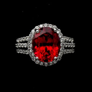 May include: A ring featuring a large, oval-cut, deep red gemstone set in a silver-toned metal band. The central stone is surrounded by a halo of small, clear, round-cut stones, with additional clear stones embedded along the triple-split band.