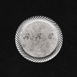 Puede incluir: Un disco circular de plata con un borde decorativo acanalado. El disco tiene las iniciales "A. A. C." grabadas en el centro.