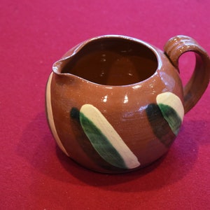 Op de afbeelding: Een bruine keramische kan met een abstract ontwerp in groen en wit. De kan heeft een rond lichaam en een handvat.