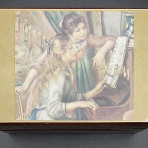 May include: A vintage wooden music box with a gold-toned frame. The lid features a painted image of two girls, one playing a piano and the other looking on. The image is reminiscent of a classic painting.