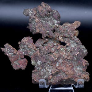 May include: A natural copper specimen with a rough, textured surface. The copper displays a mix of reddish-brown and grey-green hues, with a crystalline structure. The mineral is presented on a clear display stand against a dark background.