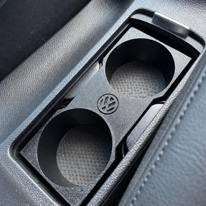 Könnte beinhalten: Ein schwarzer Doppel-Getränkehalter aus Kunststoff für die Mittelkonsole eines Fahrzeugs, mit zwei kreisförmigen Öffnungen, strukturierten Böden und einem mittig eingeprägten Volkswagen-Logo zwischen den beiden Halterungen für einen festen Sitz.