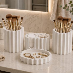 Puede incluir: Un conjunto de organizadores de baño blancos con diseño estriado que incluye dos porta brochas cilíndricos, un recipiente de dos compartimentos para discos de algodón y bastoncillos, y una bandeja circular con un reloj dorado, dos anillos y un pasador de perlas.
