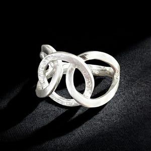 May include: A silver ring with an interlocking design. The ring features three connected, rounded shapes, creating a unique and modern look. The ring is set against a dark background, highlighting its metallic sheen.