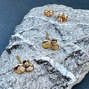Könnte beinhalten: Zwei goldene Ohrstecker, die jeweils aus drei kleinen, kreisförmigen Scheiben mit gehämmertem Muster in einer Anordnung bestehen. Das Set enthält zwei passende goldene Ohrsteckerverschlüsse, die separat oben abgebildet sind.