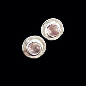 Könnte beinhalten: Ein Paar Ohrstecker mit einer kreisförmigen, gehämmerten Silbereinfassung und einem glatten, polierten, blassrosa Cabochon-Edelstein in der Mitte. Die Metalloberfläche weist eine texturierte, organische Optik rund um den runden, hellrosa Stein auf.