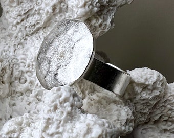 May include: A silver ring featuring a large, circular, textured face with a matte finish and a smooth, polished band. The metal surface of the circular top displays a subtle, uneven, hammered-like appearance, contrasting with the band's clean lines.