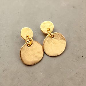 Könnte beinhalten: Ein Paar goldfarbene Ohrhänger mit zwei runden Scheiben in unterschiedlichen Größen. Jeder Ohrring besteht aus einer kleineren oberen Scheibe, die mit einem Ring an einer größeren, unregelmäßig gehämmerten unteren Scheibe befestigt ist.