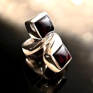 Könnte beinhalten: Ein Silberring mit zwei quadratischen, dunkelroten Edelsteinen. Der Ring hat ein modernes Design mit poliertem Finish, das Licht reflektiert. Die Edelsteine sind in einer Lünettenfassung gefasst. Schmuck.