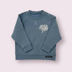 Puede incluir: Una sudadera azul pizarra con el texto "Ebbe & Flut" en escritura blanca. La parte superior de manga larga tiene cuello redondo y cierres de botón a presión en el hombro. El dobladillo inferior tiene una pequeña etiqueta negra con texto blanco.