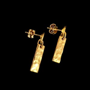 Puede incluir: Un par de pendientes dorados con un diseño rectangular de textura martillada. Los pendientes tienen un cierre de poste y mariposa. El fondo es negro liso.