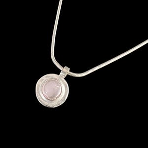 May include: A silver necklace with a round pendant. The pendant features a light pink stone set within a silver bezel. The chain is a thin, snake-style design. The background is solid black.