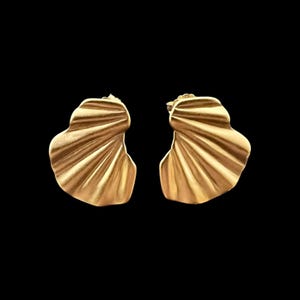 Peut inclure: Une paire de boucles d'oreilles à clous dorées présentant un design en forme de coquillage plissé avec une finition mate. Les boucles d'oreilles ont une silhouette incurvée et asymétrique avec des crêtes rayonnantes partant du fermoir.