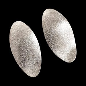 Könnte beinhalten: Ein Paar silberfarbene Ohrringe in länglicher Ovalform. Jeder Ohrring weist eine texturierte Oberfläche mit Kreuzschraffur auf, die das Licht einfängt und einen subtilen metallischen Glanz auf den gewölbten, flachen Metalloberflächen erzeugt.