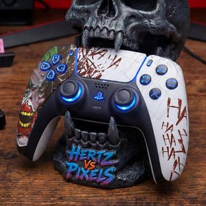 May include: A gray skull-shaped gaming controller holder with horns, holding a black and white PlayStation controller. The controller has blue lights and a graphic design with the text "HA HA HA" and "Hertz vs Pixels".