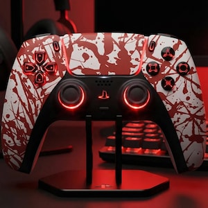 Peut inclure: Une manette de jeu personnalisée avec un design éclaboussé blanc et rouge, des boutons éclairés et une base noire. La manette est sur un support noir avec une lueur rouge, suggérant un thème de jeu.
