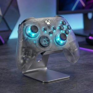 以下が含まれることがあります： 青色のLEDライトを備えた透明なXboxコントローラーが、銀色のスタンドに置かれています。コントローラーには黒いボタンと方向パッドがあります。スタンドはつや消し金属製で、背景はぼやけています。