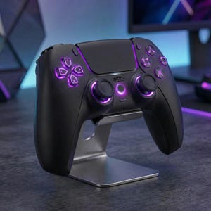 May include: A black gaming controller with purple illuminated buttons and joysticks, resting on a silver metal stand. The controller features a sleek design with a matte finish. The buttons and joysticks glow with a vibrant purple light.