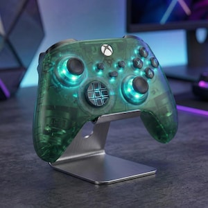 May include: A transparent green Xbox controller with illuminated buttons and a silver stand. The controller has a clear shell, revealing internal components. The buttons and thumbsticks are black. The stand is made of brushed metal.