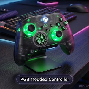 May include: A transparent gaming controller with internal green LED lighting, black thumbsticks, and a circular directional pad. The controller features the text RGB Modded Controller and is mounted on a black stand, showcasing a clear shell design.