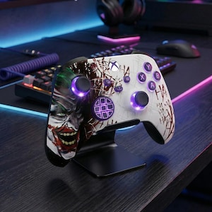May include: A white Xbox controller with a Joker-themed design, set on a black stand. The controller has purple buttons and thumbsticks, and features the Joker's face and blood splatter effects. The controller is designed for gaming.