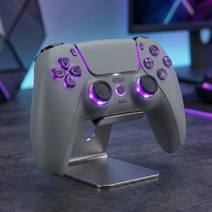 May include: A gray gaming controller with purple illuminated buttons and accents, resting on a silver metal stand. The controller features a directional pad, buttons, and analog sticks. The stand is rectangular and supports the controller at an angle.