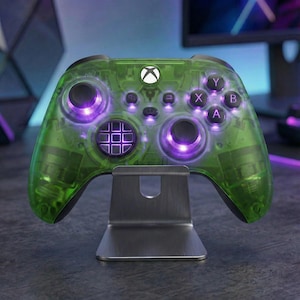 May include: A transparent green Xbox controller with purple lighting accents, resting on a silver metal stand. The buttons are labelled with white letters. The controller has a modern design.