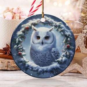 May include: A round Christmas ornament featuring a snowy owl illustration. The owl is surrounded by a wreath of greenery and red berries. The ornament hangs from a gold string, with a festive background of candy canes, hot cocoa, and other holiday decorations.