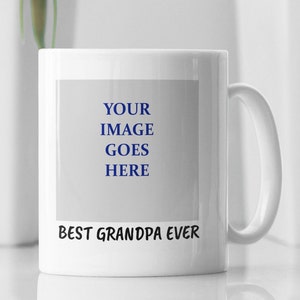 Op de afbeelding: Witte keramische mok met een groot handvat. De mok heeft de tekst "BEST GRANDPA EVER" in zwart aan de onderkant en de woorden "YOUR IMAGE GOES HERE" in blauw in een rechthoekige ruimte. De mok is ontworpen voor personalisatie.