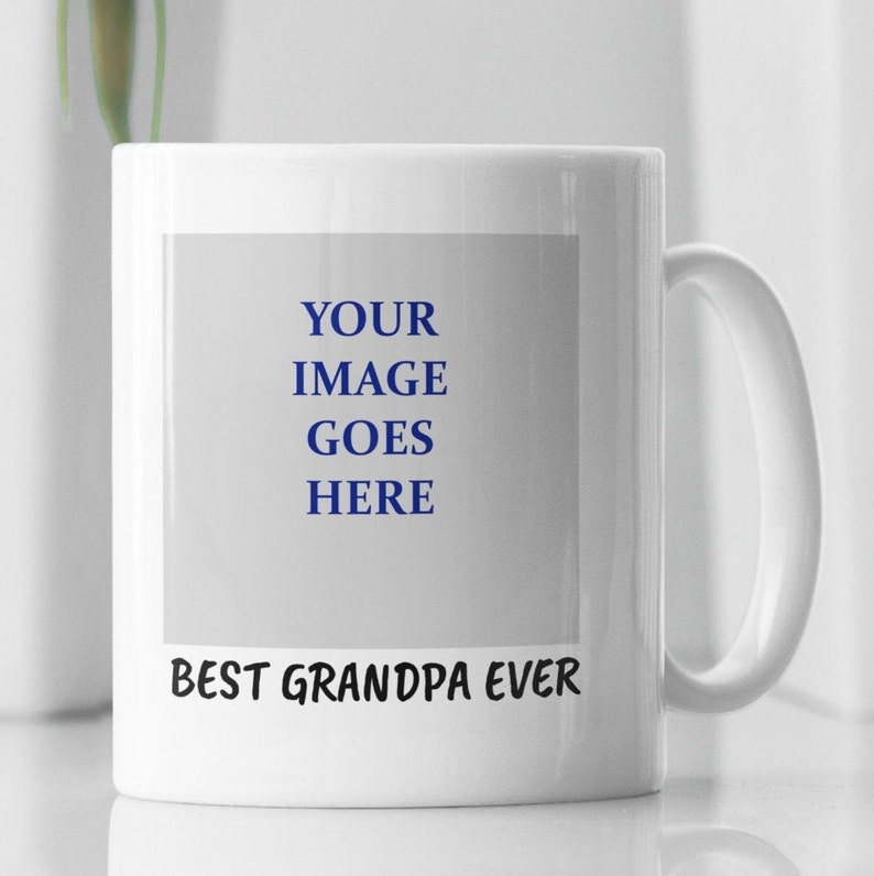 Op de afbeelding: Witte keramische mok met een groot handvat. De mok heeft de tekst "BEST GRANDPA EVER" in zwart aan de onderkant en de woorden "YOUR IMAGE GOES HERE" in blauw in een rechthoekige ruimte. De mok is ontworpen voor personalisatie.