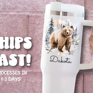 Pode incluir: Caneca t&eacute;rmica branca com uma ilustra&ccedil;&atilde;o em aquarela de um urso pardo em uma cena de floresta. O nome "Dakota" est&aacute; escrito em it&aacute;lico abaixo do urso. A caneca tem uma al&ccedil;a e uma tampa branca.