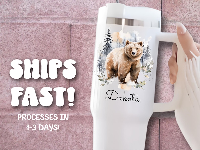 Pode incluir: Caneca t&eacute;rmica branca com uma ilustra&ccedil;&atilde;o em aquarela de um urso pardo em uma cena de floresta. O nome "Dakota" est&aacute; escrito em it&aacute;lico abaixo do urso. A caneca tem uma al&ccedil;a e uma tampa branca.