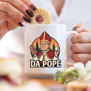 May include: White ceramic mug featuring a cartoon illustration of a pope holding a hot dog and a baseball, with the text "DA POPE". The background includes a cityscape and sunset colors.
