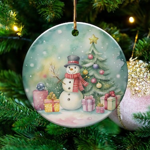 May include: A round Christmas ornament featuring a watercolor illustration of a snowman wearing a top hat and scarf, standing next to a decorated Christmas tree and wrapped gifts. The ornament is hanging on a Christmas tree.