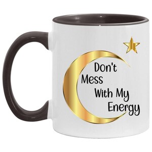 Puede incluir: Taza de cer&aacute;mica blanca con asa y borde marr&oacute;n oscuro. La taza presenta un dise&ntilde;o de luna creciente y estrella doradas con el texto "Don't Mess With My Energy" en negro. Una taza de caf&eacute; elegante y motivadora.