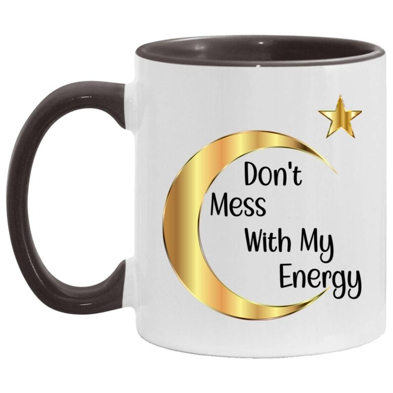 Puede incluir: Taza de cer&aacute;mica blanca con asa y borde marr&oacute;n oscuro. La taza presenta un dise&ntilde;o de luna creciente y estrella doradas con el texto "Don't Mess With My Energy" en negro. Una taza de caf&eacute; elegante y motivadora.