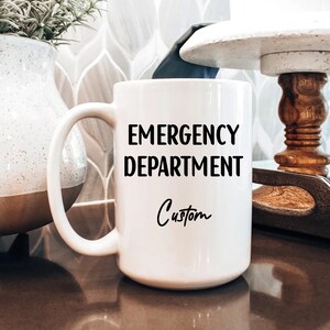 Pu&ograve; includere: Una tazza di ceramica bianca con un manico grande. La tazza presenta le parole "EMERGENCY DEPARTMENT" in grassetto nero, con "Custom" scritto sotto in un carattere corsivo. La tazza &egrave; su una superficie marrone.