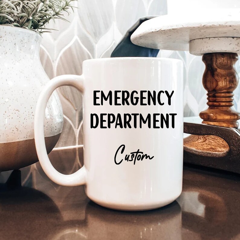 Pu&ograve; includere: Una tazza di ceramica bianca con un manico grande. La tazza presenta le parole "EMERGENCY DEPARTMENT" in grassetto nero, con "Custom" scritto sotto in un carattere corsivo. La tazza &egrave; su una superficie marrone.