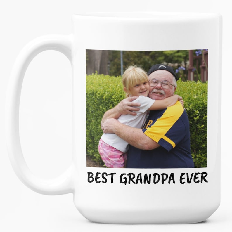 Op de afbeelding: Witte keramische mok met een foto van een lachende man en een jong meisje dat elkaar omhelst. De tekst "BEST GRANDPA EVER" staat onder de foto. De mok heeft een groot handvat.