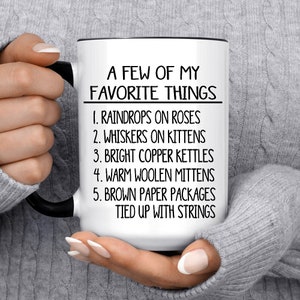 May include: White ceramic mug with a black rim and handle. The mug has black text that reads "A Few of My Favorite Things" followed by a list: "1. Raindrops on Roses, 2. Whiskers on Kittens, 3. Bright Copper Kettles, 4. Warm Woolen Mittens, 5. Brown Paper Packages Tied Up With Strings."