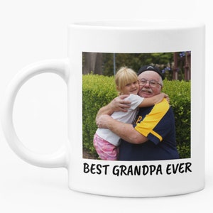 Op de afbeelding: Witte keramische mok met een foto van een opa die een jong meisje omhelst. De afbeelding staat tegen een groene achtergrond. De tekst "BEST GRANDPA EVER" is in het zwart op de mok gedrukt.
