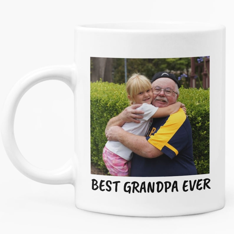Op de afbeelding: Witte keramische mok met een foto van een opa die een jong meisje omhelst. De afbeelding staat tegen een groene achtergrond. De tekst "BEST GRANDPA EVER" is in het zwart op de mok gedrukt.