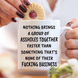 May include: White ceramic mug with the text "NOTHING BRINGS A GROUP OF ASSHOLES TOGETHER FASTER THAN SOMETHING THAT'S NONE OF THEIR FUCKING BUSINESS" in black. A cookie with a red center sits on top of the mug.