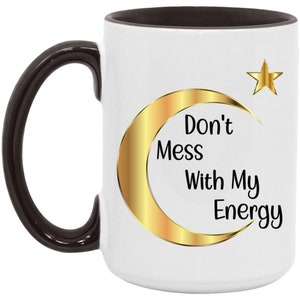 Puede incluir: Taza de cer&aacute;mica blanca con asa e interior negros. La taza presenta un dise&ntilde;o de luna creciente y estrella doradas con el texto "Don't Mess With My Energy" en negro. Una taza de caf&eacute; elegante y motivadora.