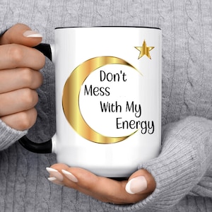 Puede incluir: Taza de cer&aacute;mica blanca con interior y asa negros. La taza presenta un dise&ntilde;o de media luna y estrella doradas con el texto "Don't Mess With My Energy" en negro. Un regalo elegante y motivador.