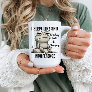 May include: White ceramic mug with a black and white illustration of a frog sitting on a rock. The mug has the text "I slept like shit so today I will be choosing indifference" printed on it.