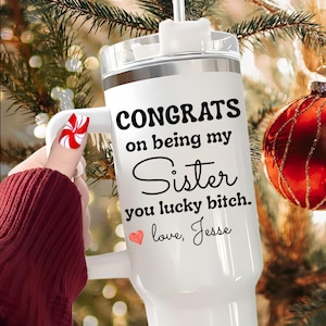 May include: White insulated tumbler with a handle and a lid. The tumbler has the text "CONGRATS on being my Sister you lucky bitch. love, Jesse" in black script. A red heart graphic is also present.