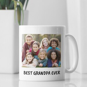 Op de afbeelding: Witte keramische mok met een foto van zeven kinderen en de tekst "BEST GRANDPA EVER" in het zwart. De kinderen lachen en liggen op het gras. De mok heeft een wit oor en staat op een wit oppervlak.